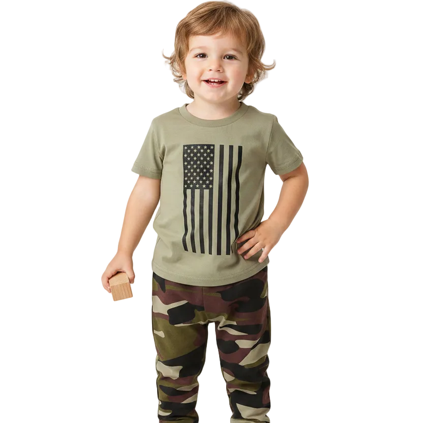 A smiling toddler standing barefoot against a clean white studio background, wearing an olive green short-sleeve T-shirt with a black American flag graphic on the front and camouflage-print pants.