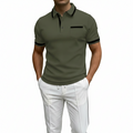 A man wearing a fitted short-sleeve polo shirt in olive green with black trim on the collar, placket, sleeve cuffs, and chest pocket, paired with white pants against a warm brown gradient background.