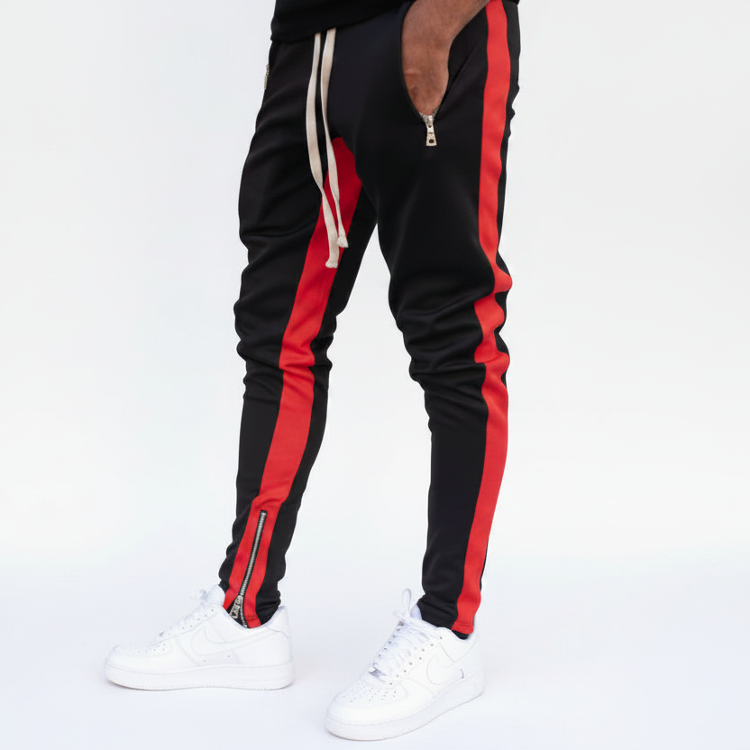 Black track pants with bold red side stripes, white drawstrings, and zippered ankles, shown on a model wearing white sneakers against a white studio background.