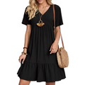 A woman wearing a black short-sleeve V-neck pleated dress with a tiered hem, styled with a woven straw shoulder bag and jewelry, standing against a textured white wall.